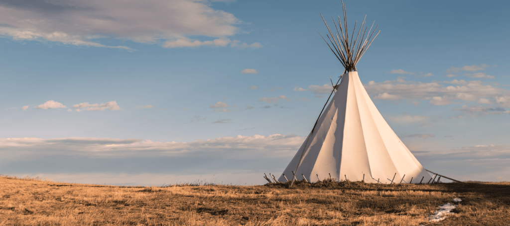 A photo of a tipi in the prairie with a blue sky with clouds in the background.